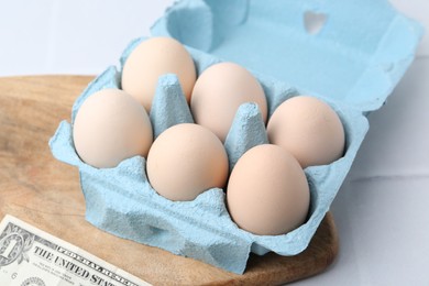 Photo of Raw chicken eggs in egg carton and dollar banknote on white tiled table, closeup