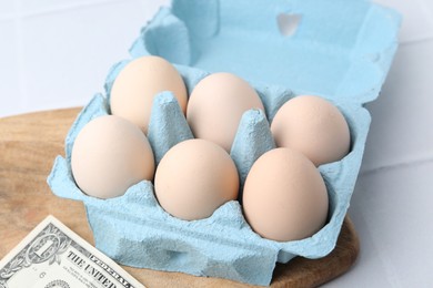 Photo of Raw chicken eggs in egg carton and dollar banknote on white tiled table, closeup
