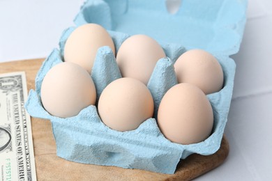 Photo of Raw chicken eggs in egg carton and dollar banknote on white tiled table, closeup