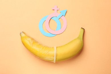 Photo of Sex education. Banana with condom, paper female and male gender symbols on pale orange background, flat lay