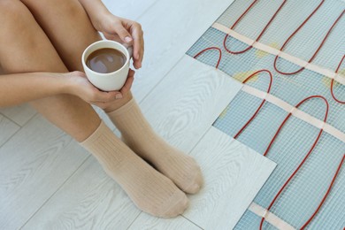 Image of Woman with coffee sitting on warm floor, closeup. Underfloor heating system