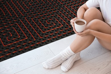 Image of Woman with coffee sitting on warm floor indoors, closeup. Underfloor heating system illustration