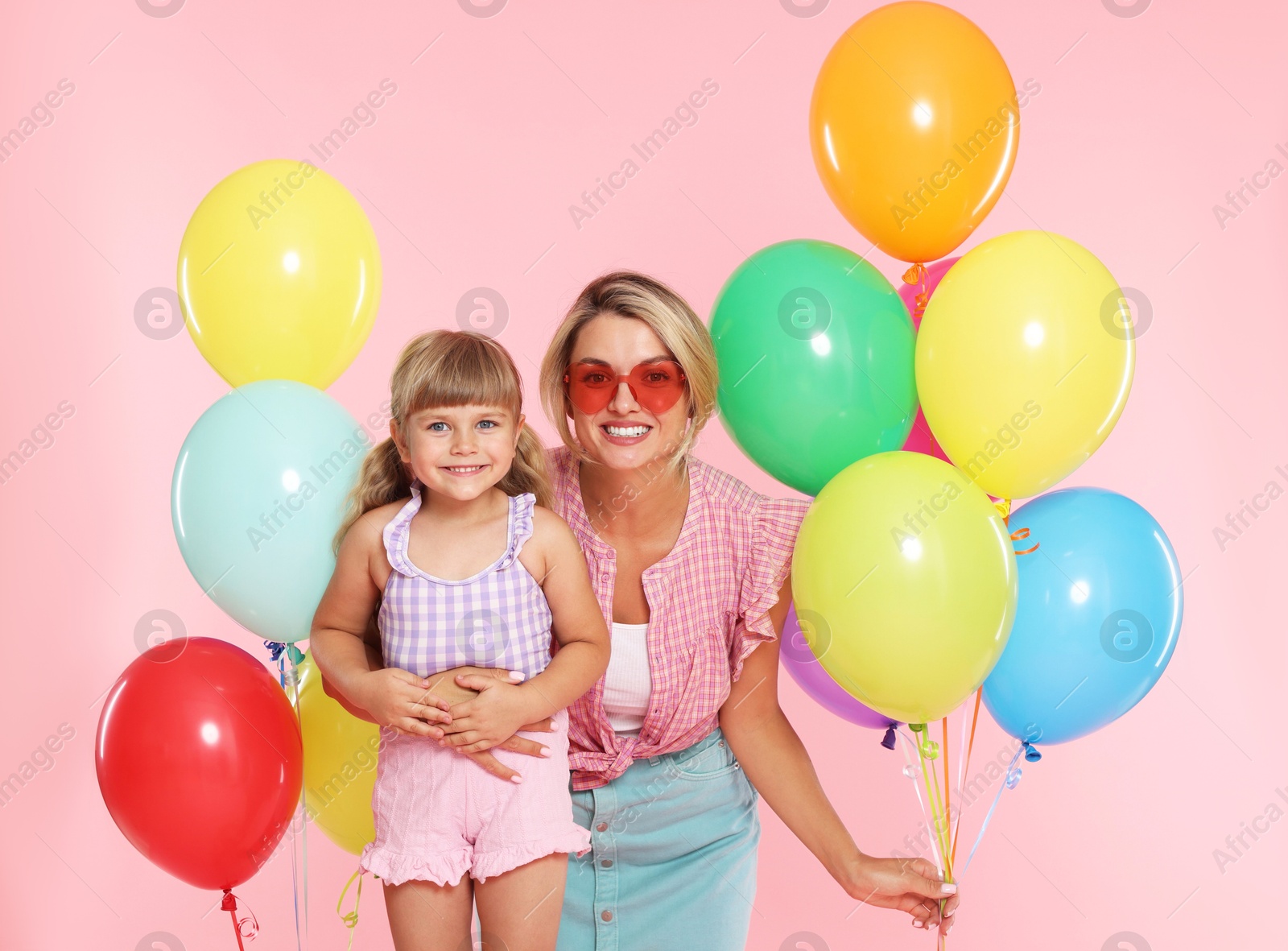 Photo of Happy mother in sunglasses and her little daughter with bunches of colorful balloons on pink background