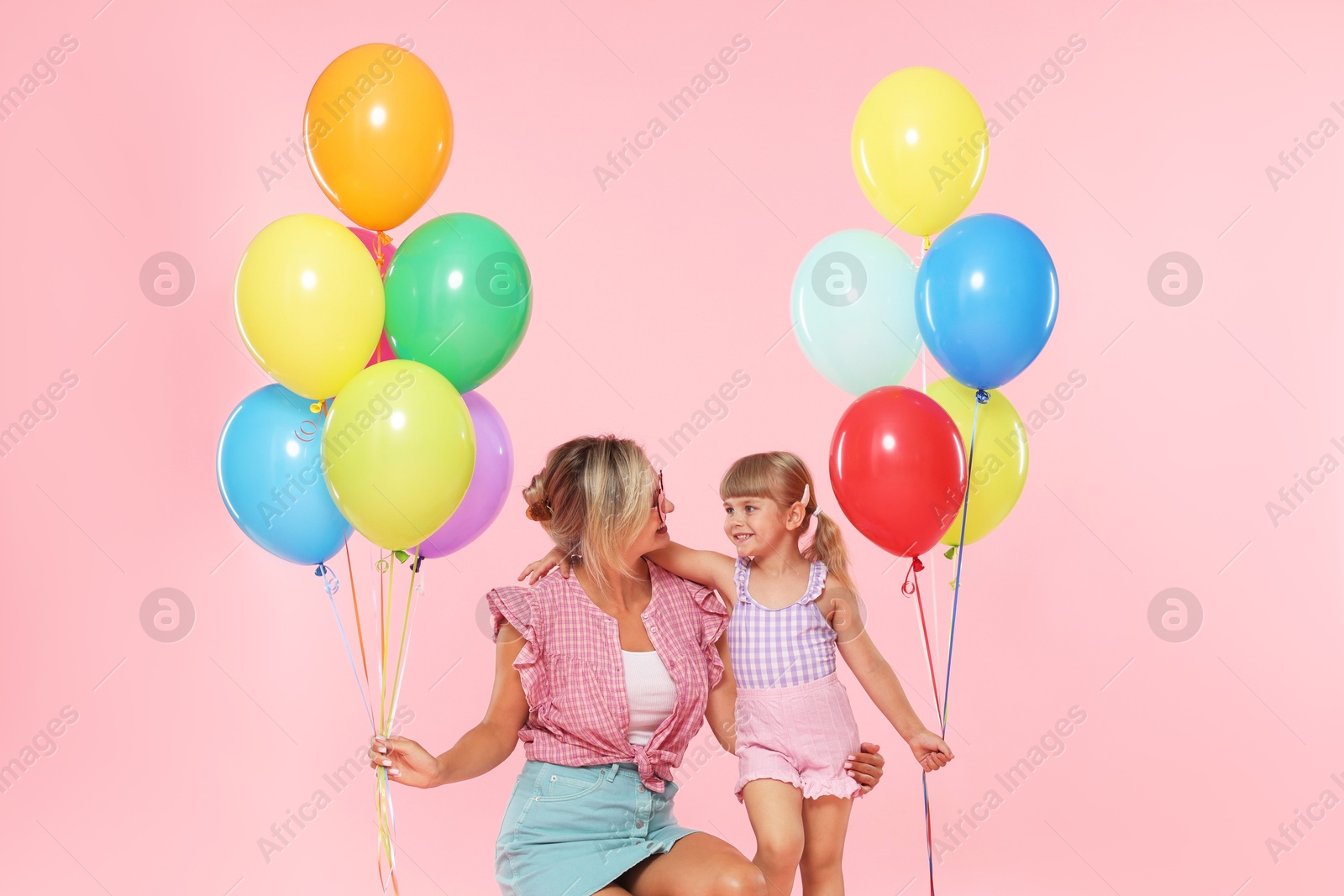 Photo of Happy mother in sunglasses and her little daughter with bunches of colorful balloons on pink background