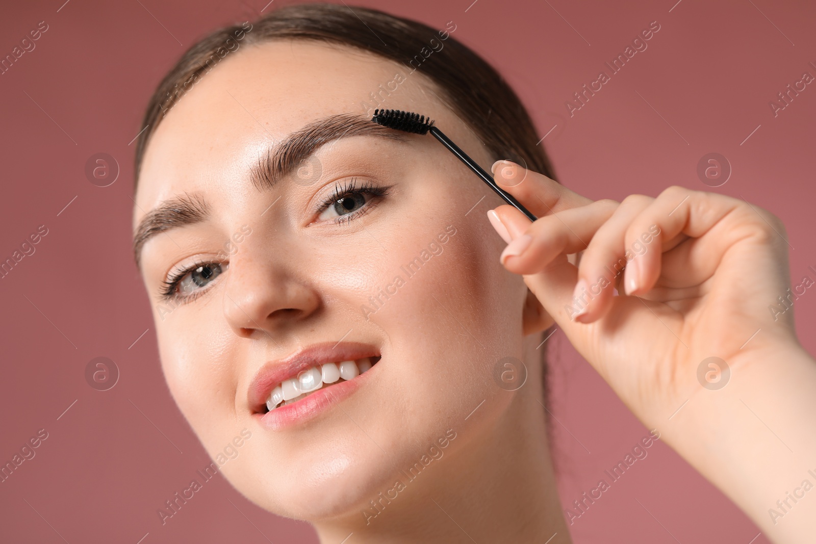 Photo of Young woman brushing her eyebrow with spoolie on pink background, closeup