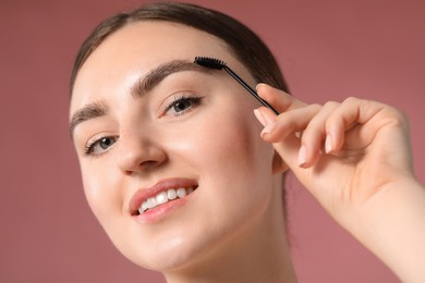 Photo of Young woman brushing her eyebrow with spoolie on pink background, closeup