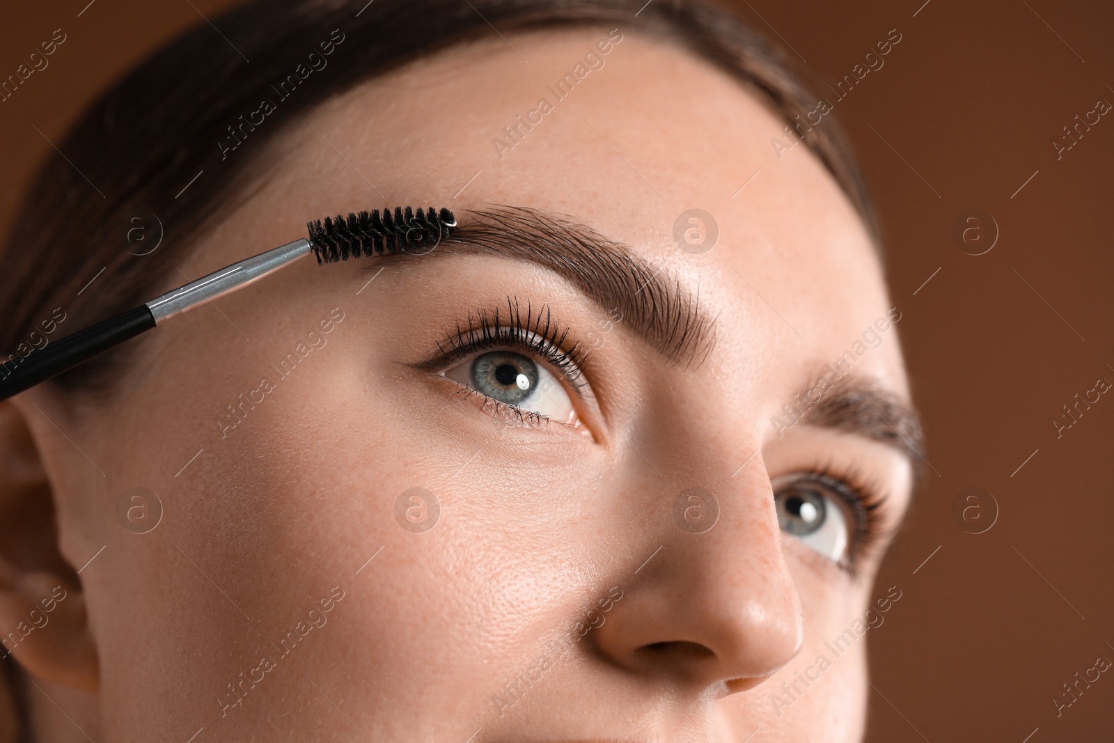 Photo of Young woman brushing her eyebrow with spoolie on brown background, closeup