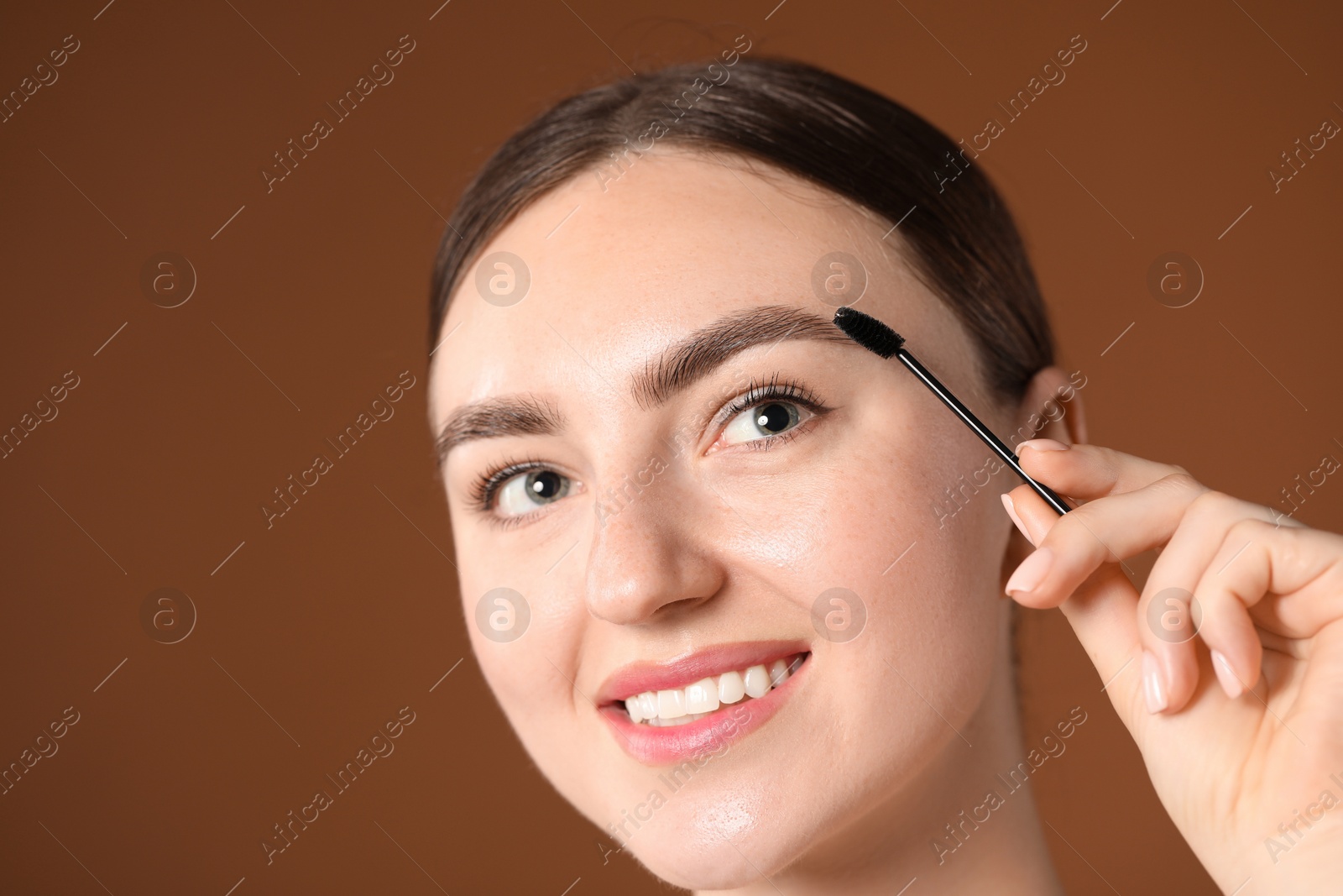 Photo of Young woman brushing her eyebrow with spoolie on brown background, closeup