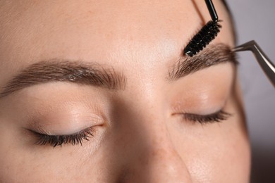 Photo of Plucking and brushing young woman's eyebrow with spoolie on light grey background, closeup
