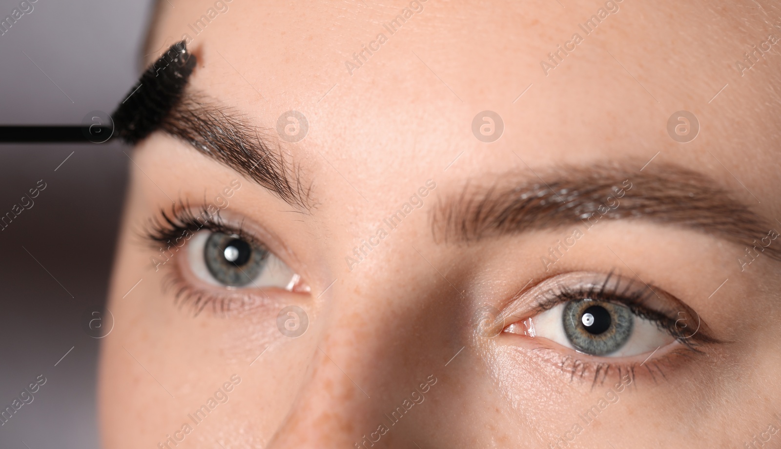 Young woman brushing her eyebrow with spoolie on light grey background, closeup Photo of Young woman brushing her eyebrow with spoolie on light grey background, closeup