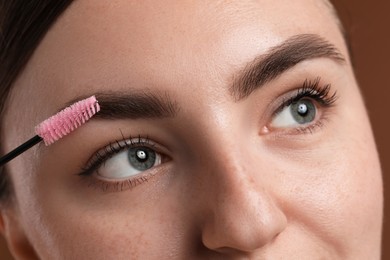 Photo of Young woman brushing her eyebrow with spoolie on brown background, closeup