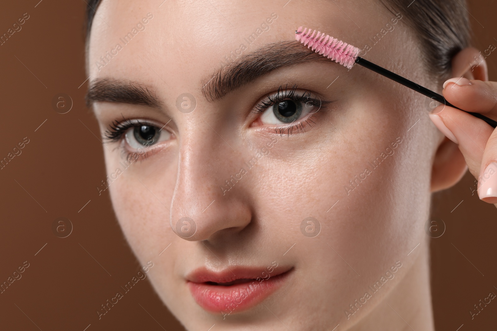 Photo of Young woman brushing her eyebrow with spoolie on brown background, closeup