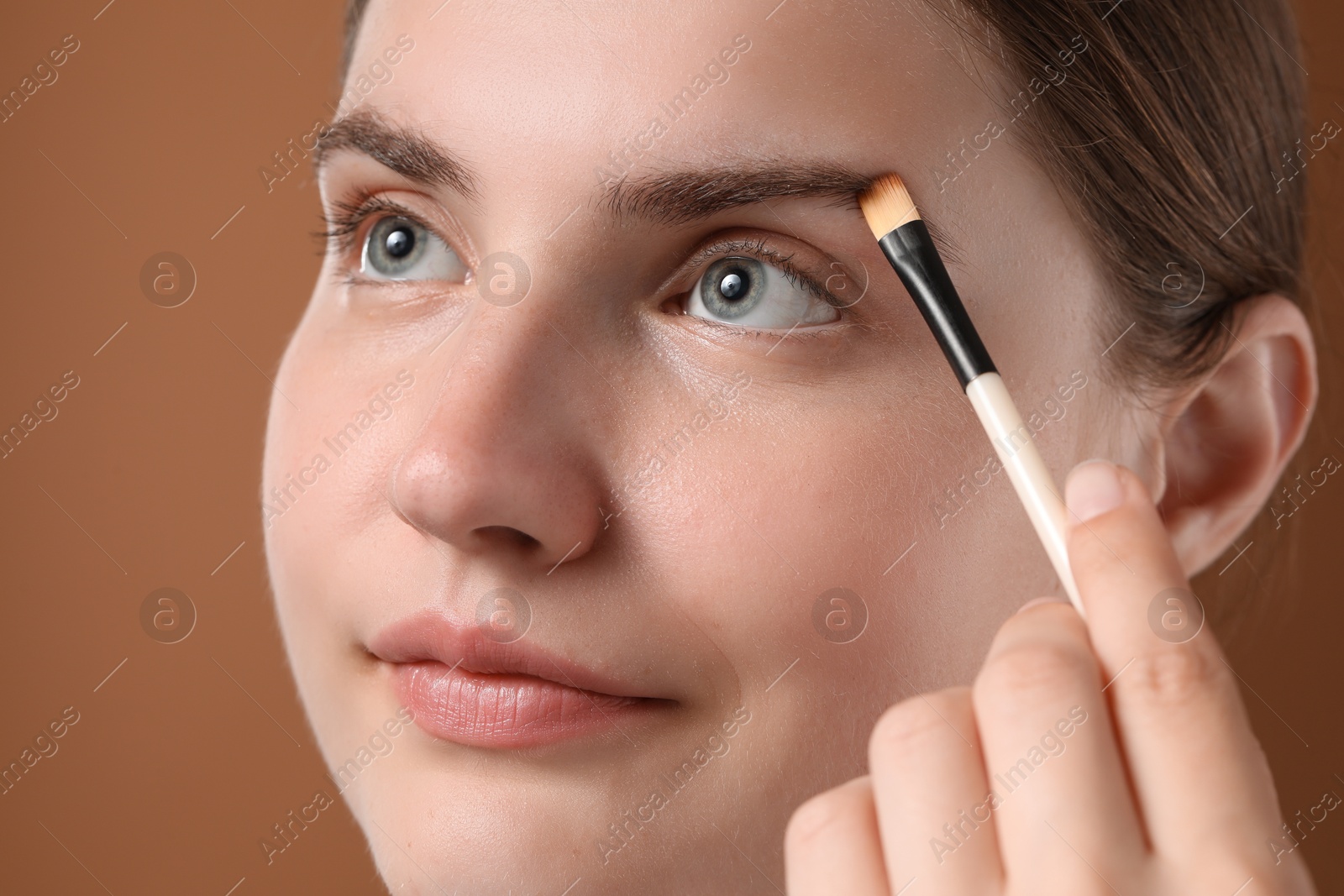 Photo of Young woman applying cosmetic product onto her eyebrow with brush on brown background, closeup