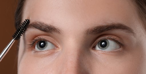 Photo of Young woman brushing her eyebrow with spoolie on brown background, closeup
