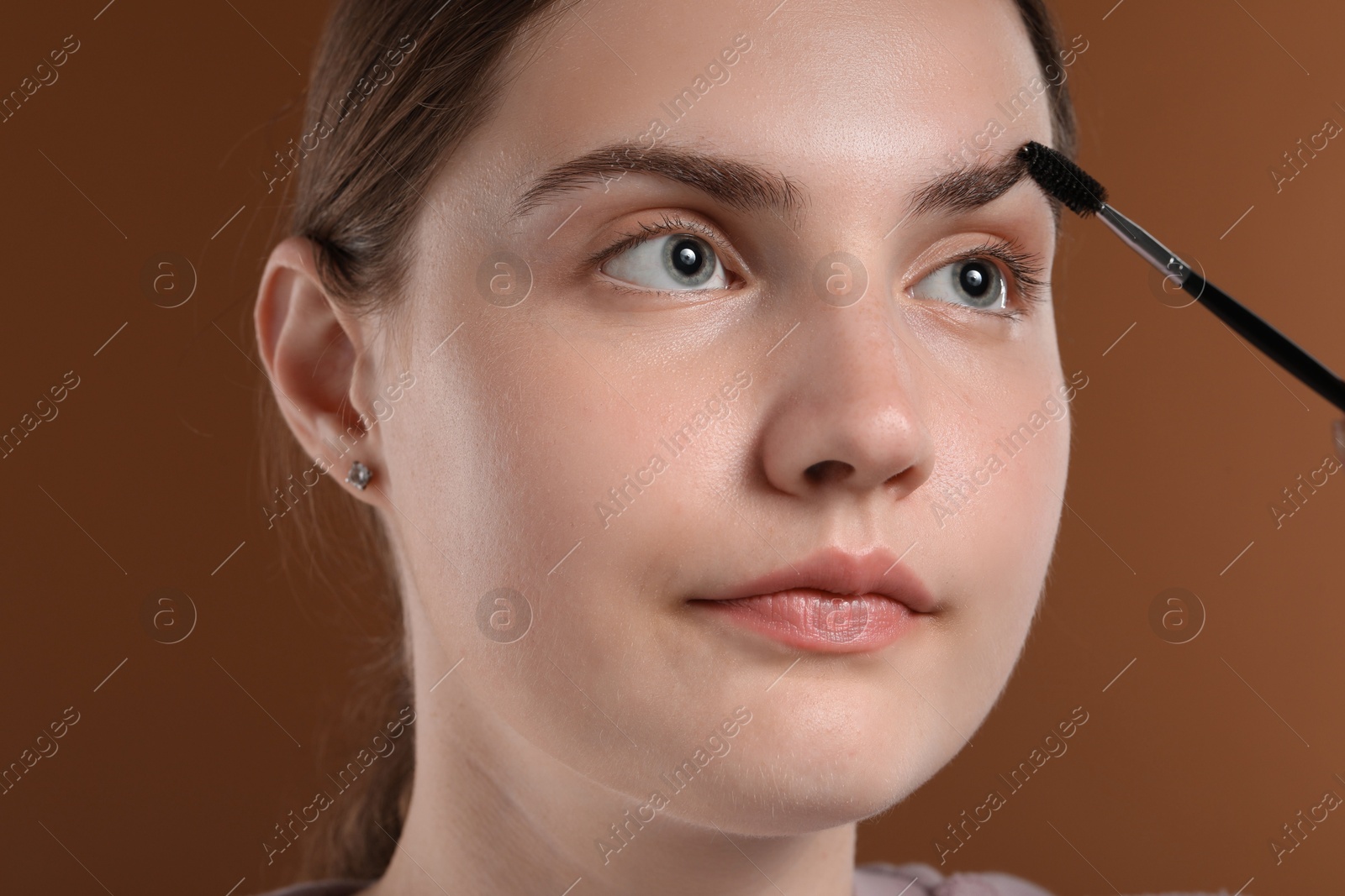 Photo of Young woman brushing her eyebrow with spoolie on brown background, closeup