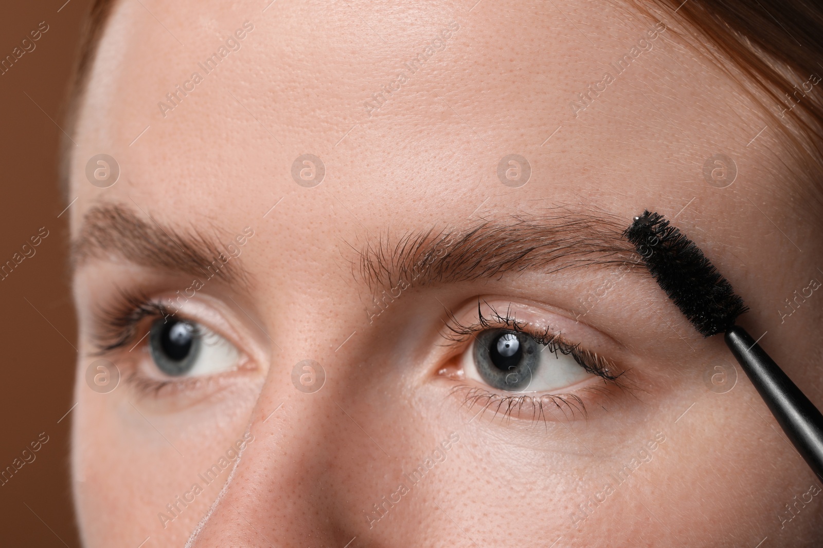 Photo of Young woman brushing her eyebrow with spoolie on brown background, closeup