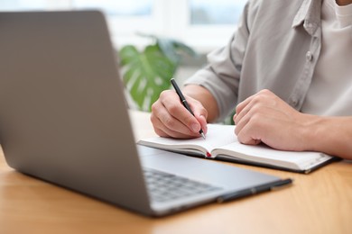 Photo of Copywriter using laptop and writing in notebook at wooden table indoors, closeup