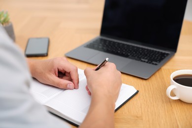 Photo of Copywriter with laptop writing in notebook at wooden table indoors, closeup