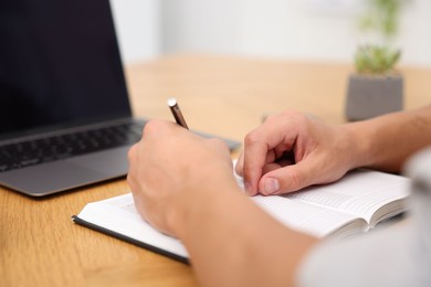 Photo of Copywriter with laptop writing in notebook at wooden table indoors, closeup