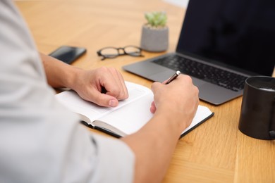 Photo of Copywriter with laptop writing in notebook at wooden table indoors, closeup