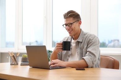 Photo of Copywriter with cup of drink using laptop at wooden table indoors