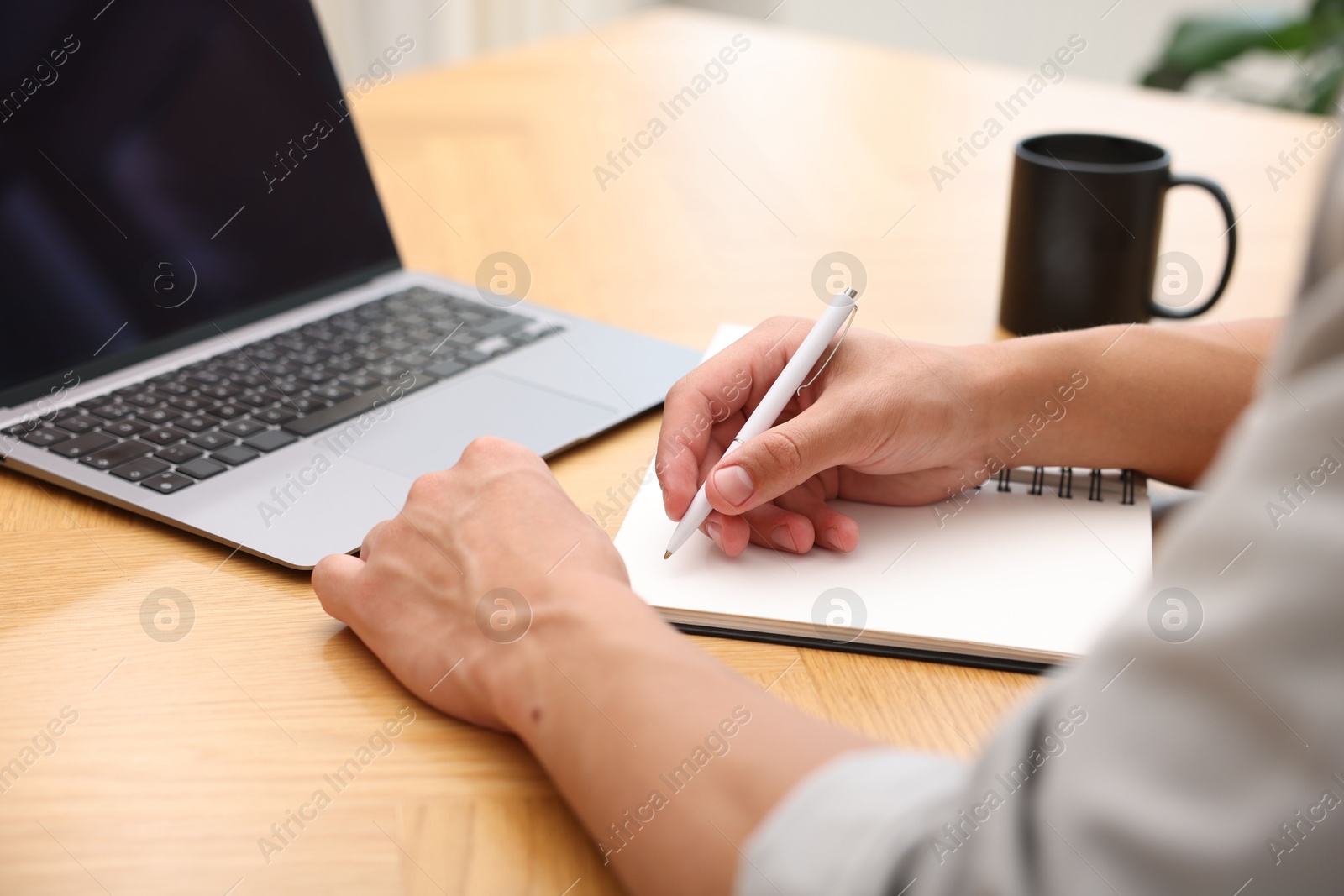 Copywriter with laptop writing in notebook at wooden table indoors, closeup Photo of Copywriter with laptop writing in notebook at wooden table indoors, closeup