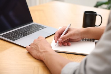 Photo of Copywriter with laptop writing in notebook at wooden table indoors, closeup