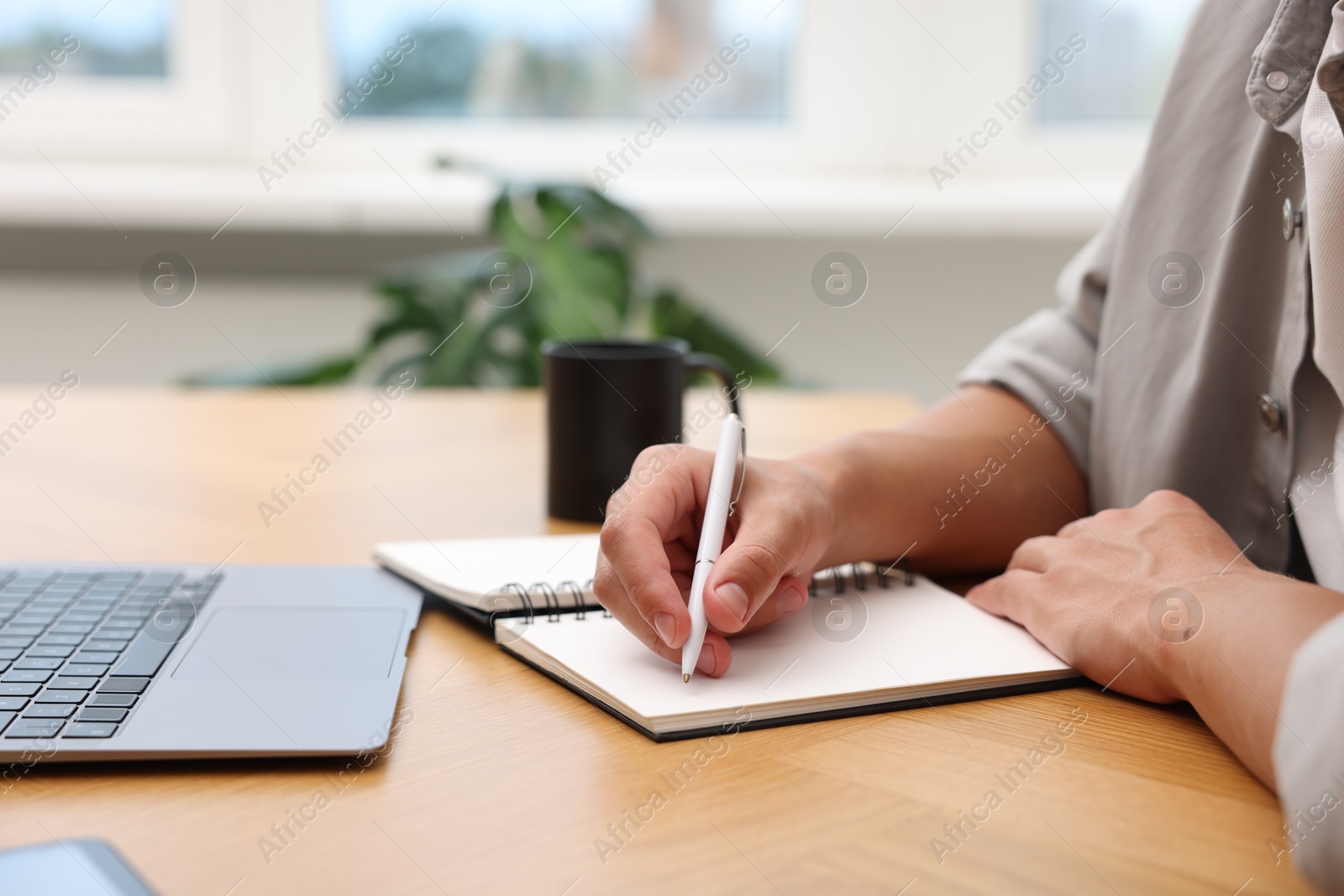 Copywriter with laptop writing in notebook at wooden table indoors, closeup Photo of Copywriter with laptop writing in notebook at wooden table indoors, closeup