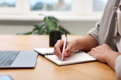 Photo of Copywriter with laptop writing in notebook at wooden table indoors, closeup