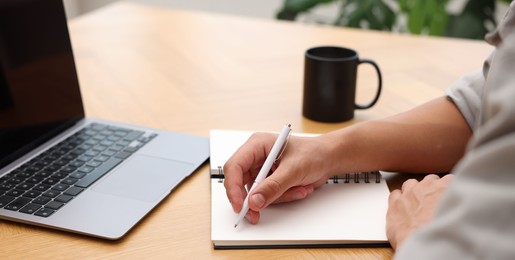 Photo of Copywriter with laptop writing in notebook at wooden table indoors, closeup