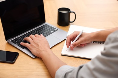 Photo of Copywriter using laptop and writing in notebook at wooden table indoors, closeup