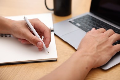 Photo of Copywriter using laptop and writing in notebook at wooden table indoors, closeup