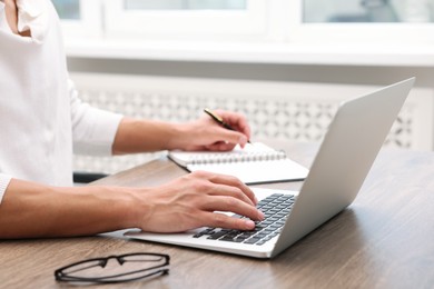Photo of Copywriter using laptop and writing in notebook at wooden table indoors, closeup