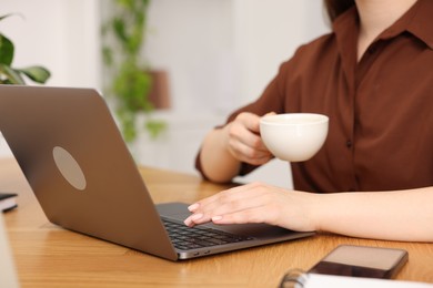 Photo of Copywriter with cup of drink using laptop at wooden table indoors, closeup