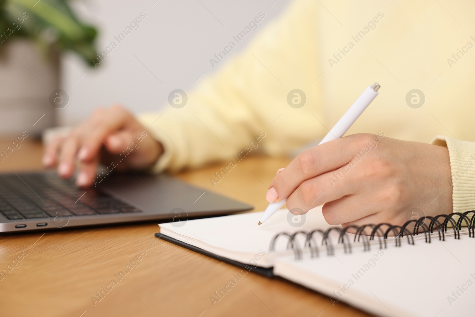 Photo of Copywriter using laptop and writing in notebook at wooden table indoors, closeup