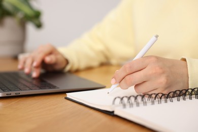 Photo of Copywriter using laptop and writing in notebook at wooden table indoors, closeup