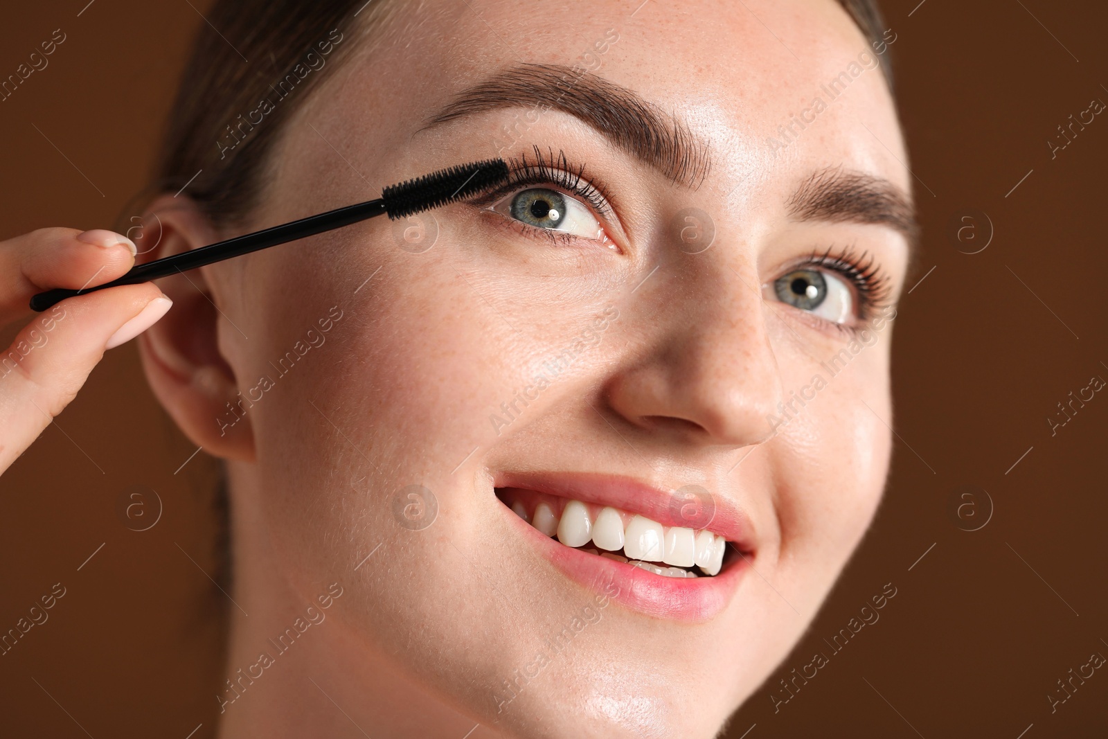 Young woman applying mascara onto her eyelashes on brown background, closeup Photo of Young woman applying mascara onto her eyelashes on brown background, closeup