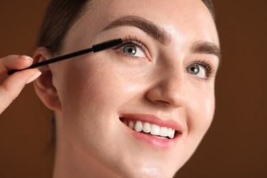Photo of Young woman applying mascara onto her eyelashes on brown background, closeup
