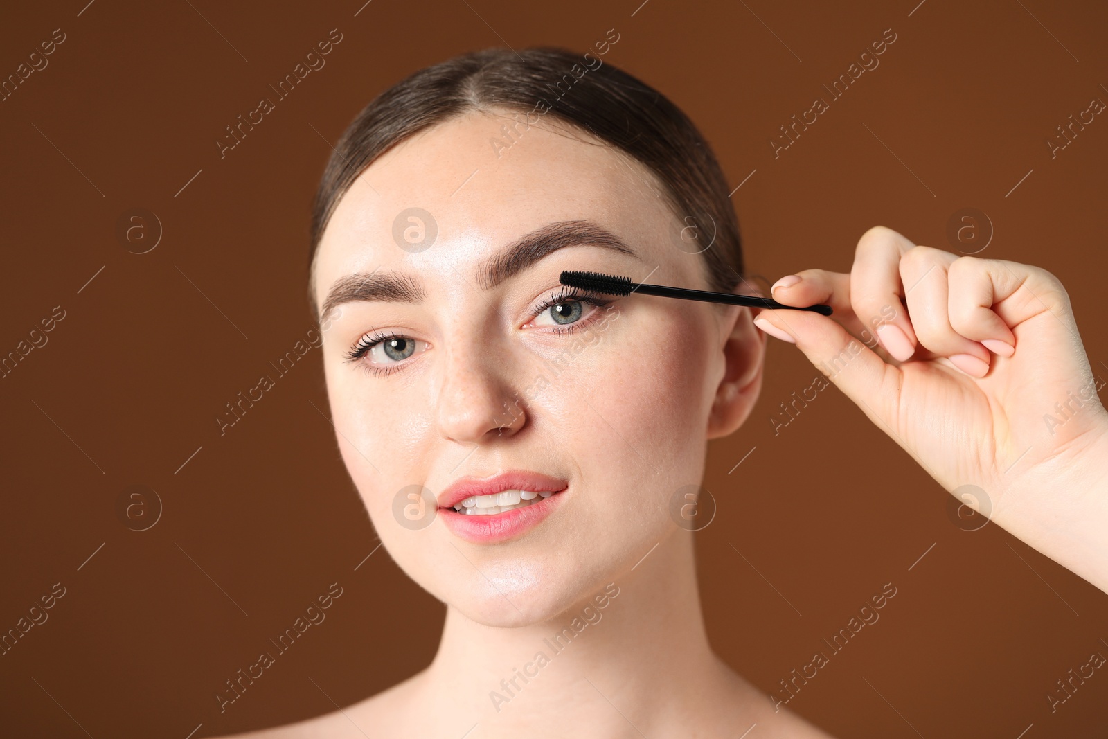 Young woman applying mascara onto her eyelashes on brown background Photo of Young woman applying mascara onto her eyelashes on brown background
