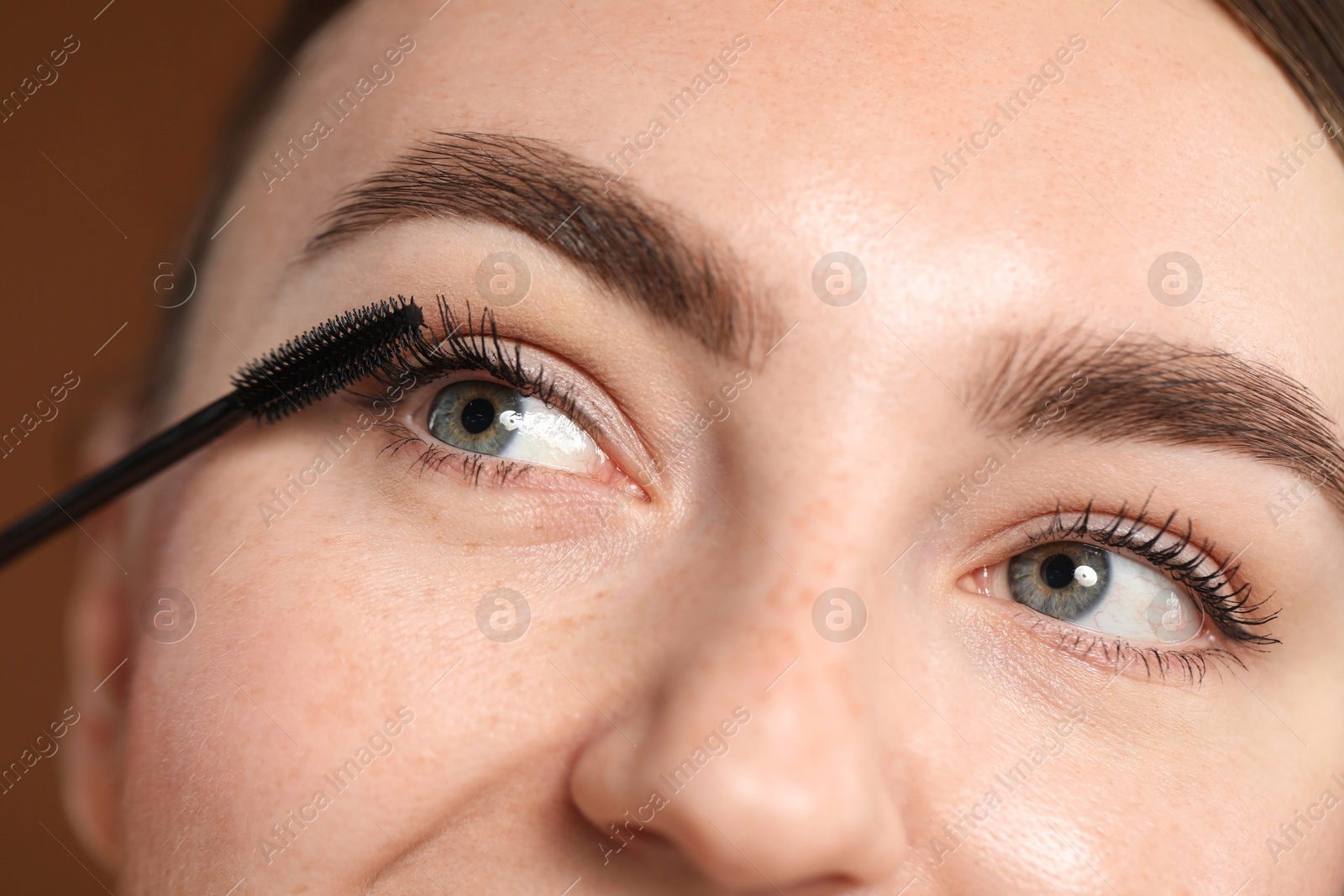 Young woman applying mascara onto her eyelashes on brown background, closeup Photo of Young woman applying mascara onto her eyelashes on brown background, closeup