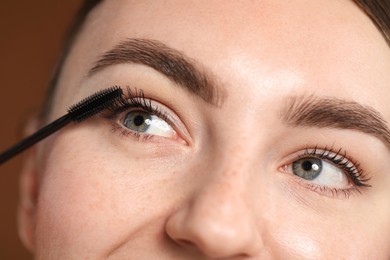 Photo of Young woman applying mascara onto her eyelashes on brown background, closeup