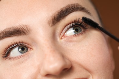 Photo of Young woman applying mascara onto her eyelashes on brown background, closeup