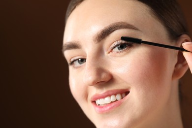 Photo of Young woman applying mascara onto her eyelashes on brown background, closeup. Space for text