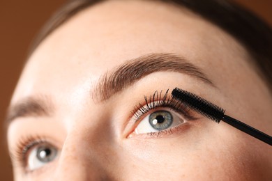 Photo of Young woman applying mascara onto her eyelashes on brown background, closeup