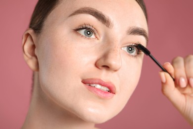 Photo of Young woman applying mascara onto her eyelashes on pink background, closeup