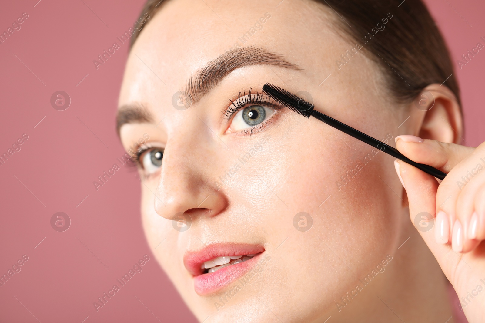 Young woman applying mascara onto her eyelashes on pink background, closeup Photo of Young woman applying mascara onto her eyelashes on pink background, closeup
