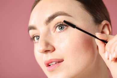 Photo of Young woman applying mascara onto her eyelashes on pink background, closeup