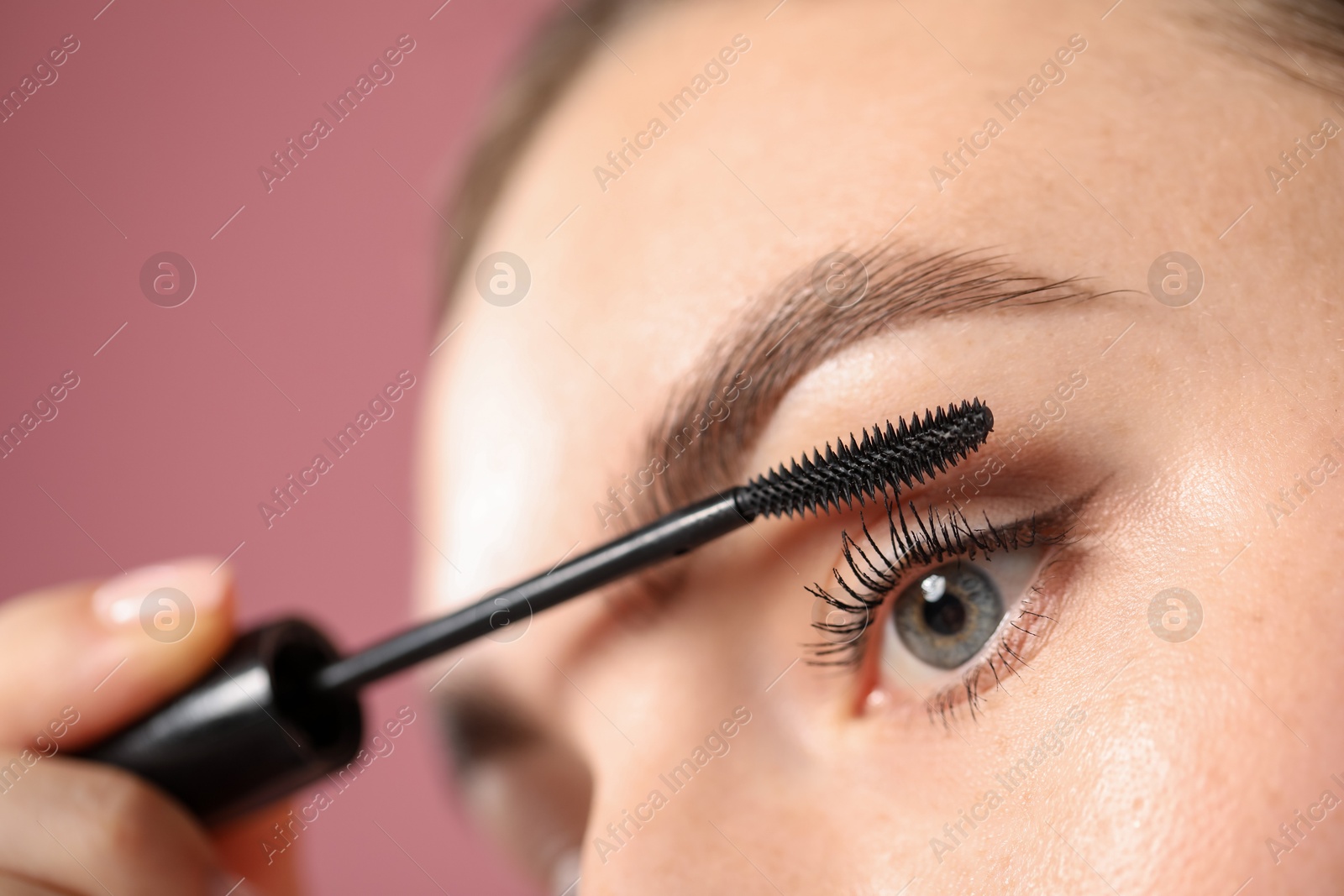 Young woman applying mascara onto her eyelashes on pink background, closeup Photo of Young woman applying mascara onto her eyelashes on pink background, closeup