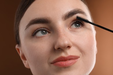 Photo of Young woman applying mascara onto her eyelashes on brown background, closeup