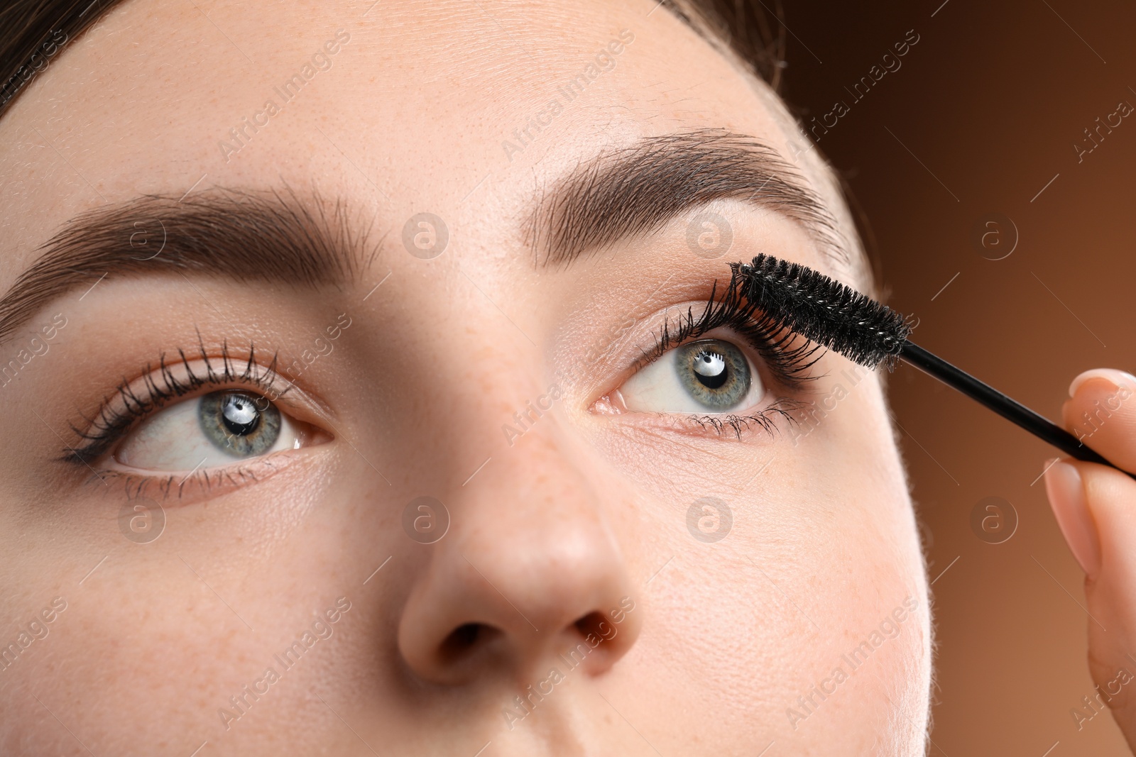 Young woman applying mascara onto her eyelashes on brown background, closeup Photo of Young woman applying mascara onto her eyelashes on brown background, closeup